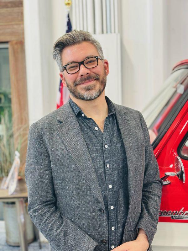 Headshot of Danny, a middle-aged, light-skinned man with salt-and-pepper hair and a cropped salt-and-pepper beard. He is wearing black glasses, a gray button-up shirt, and a gray blazer.