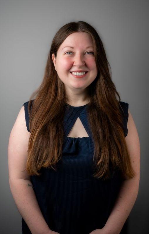 Headshot of Coral, a young Caucasian woman with long brown hair and blue eyes. She is wearing a blue sleeveless blouse.