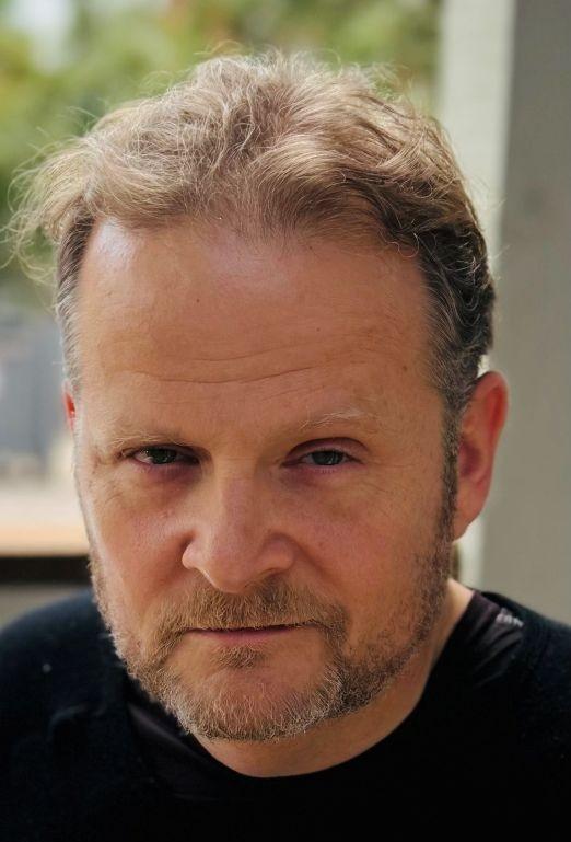 Headshot of Ben, a middle-aged Caucasian man with light, short, wavy hair and a cropped salt-and-pepper beard. He is wearing a black crew neck shirt.